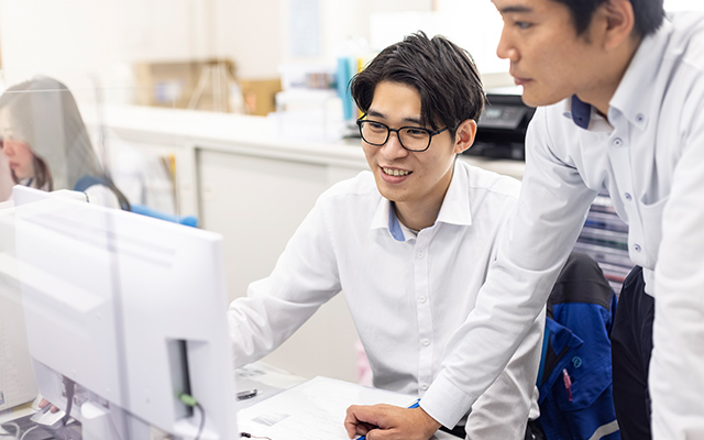 机に座っている男性社員と、その隣に立っている男性社員がパソコンを見ている様子